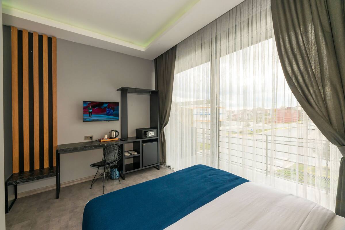 A hotel room with a bed, a desk with a chair, a television, and a view of the city through large windows.