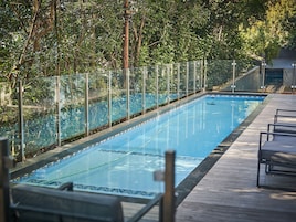A swimming pool with a wooden deck and glass railings, surrounded by trees.