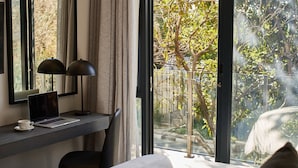 Ein Hotelzimmer mit Schreibtisch, Laptop und Tasse. Ein Bett mit weißer Bettdecke und ein Fenster mit Blick auf Bäume und einen Zaun.