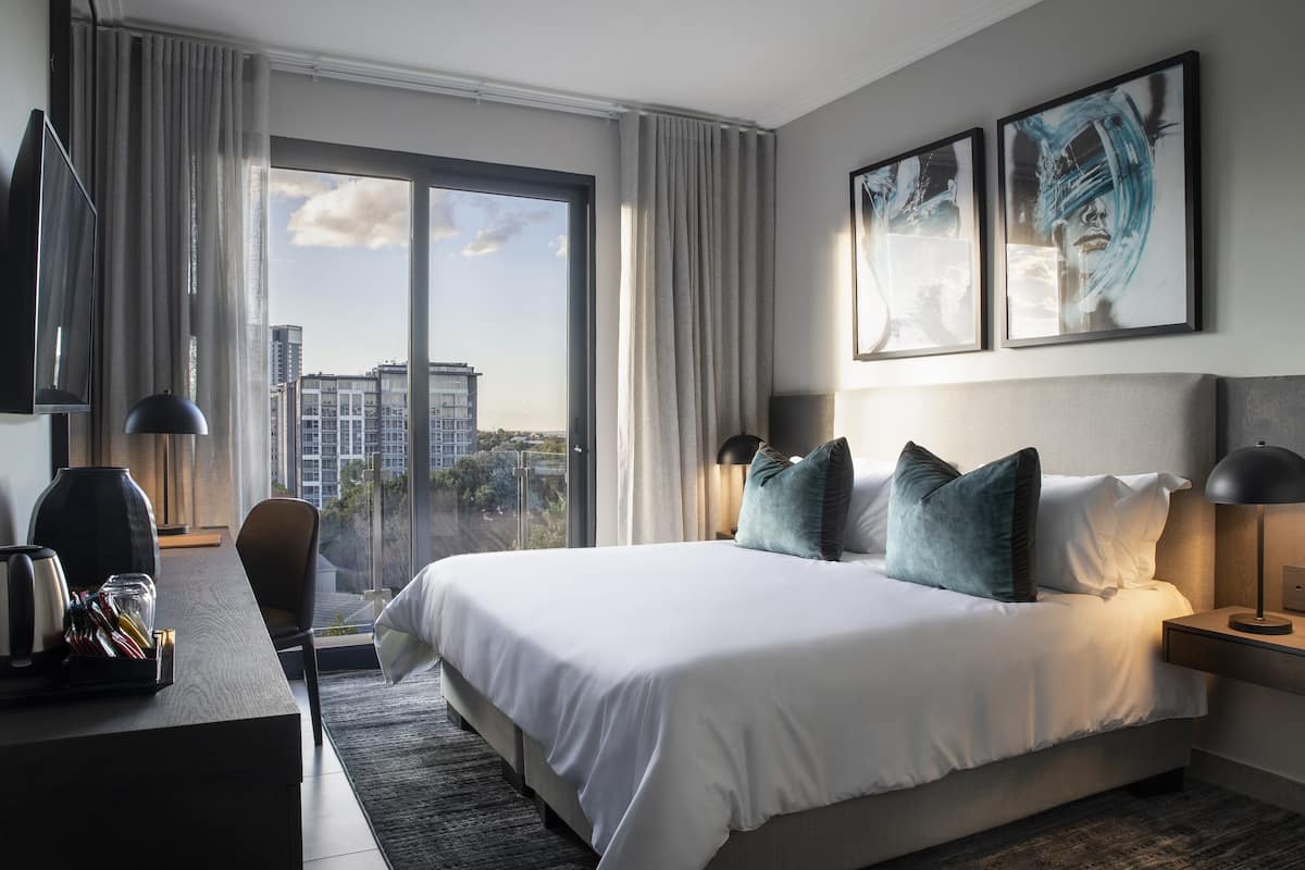 Una habitación de hotel moderna con una cama grande, un escritorio con lámpara y vistas a la ciudad a través de amplias ventanas.