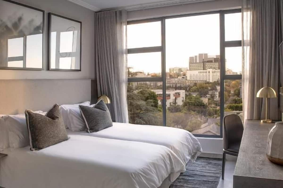 Habitación de hotel con dos camas, un ventanal amplio con vista a la ciudad, un televisor de pantalla plana y un escritorio con lámpara.