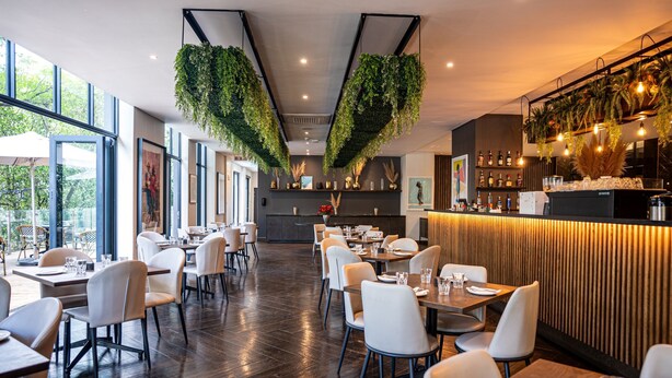 Ein modernes Restaurant mit hohen Decken, hängenden Pflanzen und einer Bar mit einer Wand in Holzoptik.