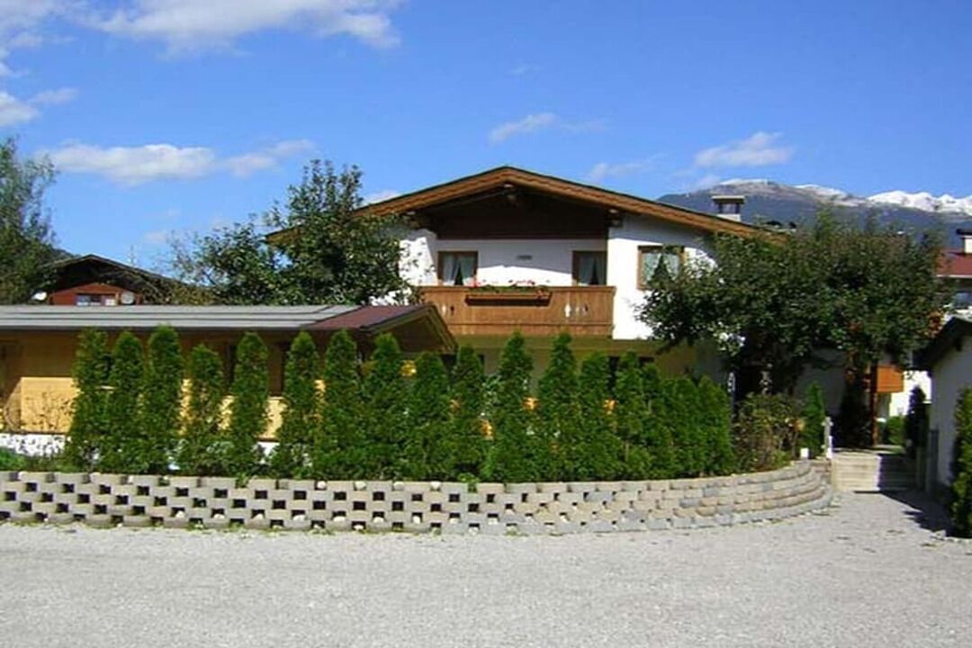 A two-story chalet with a wooden balcony, surrounded by a hedge and a stone wall.