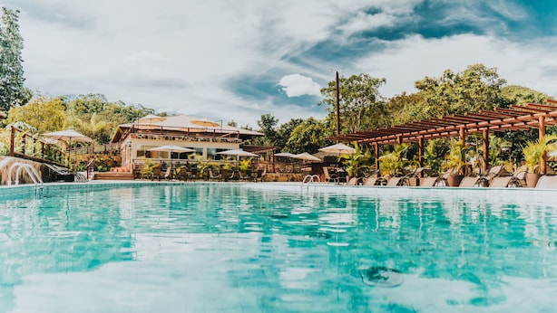 Zona de piscina con sillones reclinables y sombrillas, un edificio de varios niveles con asientos al aire libre, rodeado de árboles y un cielo despejado.