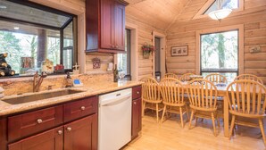 A wooden cabin interior with a kitchen area featuring a granite countertop, wooden cabinets, a dishwasher, and a dining table with wooden chairs. - Third night free on stays before 9/8/26 (Rhododendron)