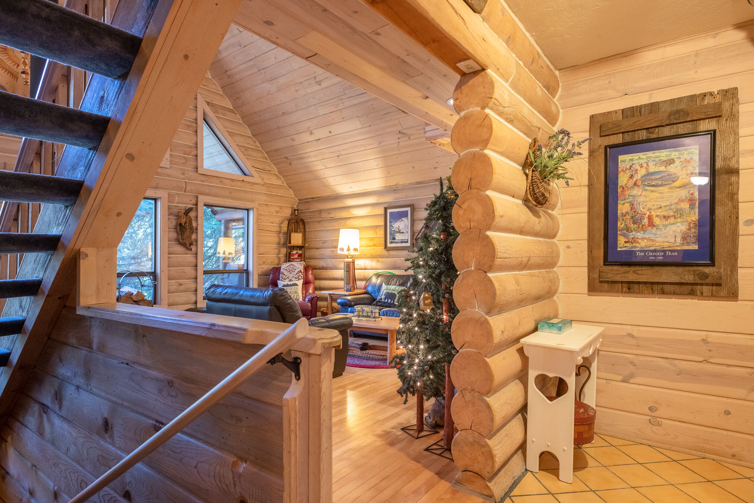 A log cabin interior with a staircase, a Christmas tree, a framed picture, and a cozy seating area.