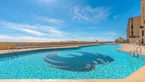 Una piscina azul cristalina con un diseño circular en el centro, rodeada de tumbonas y un edificio con balcones.