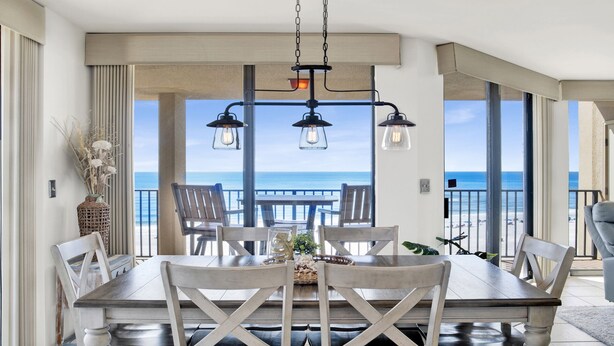 Un área de comedor con vista al océano, que incluye una mesa y sillas de madera, lámparas colgantes y un jarrón con flores secas.