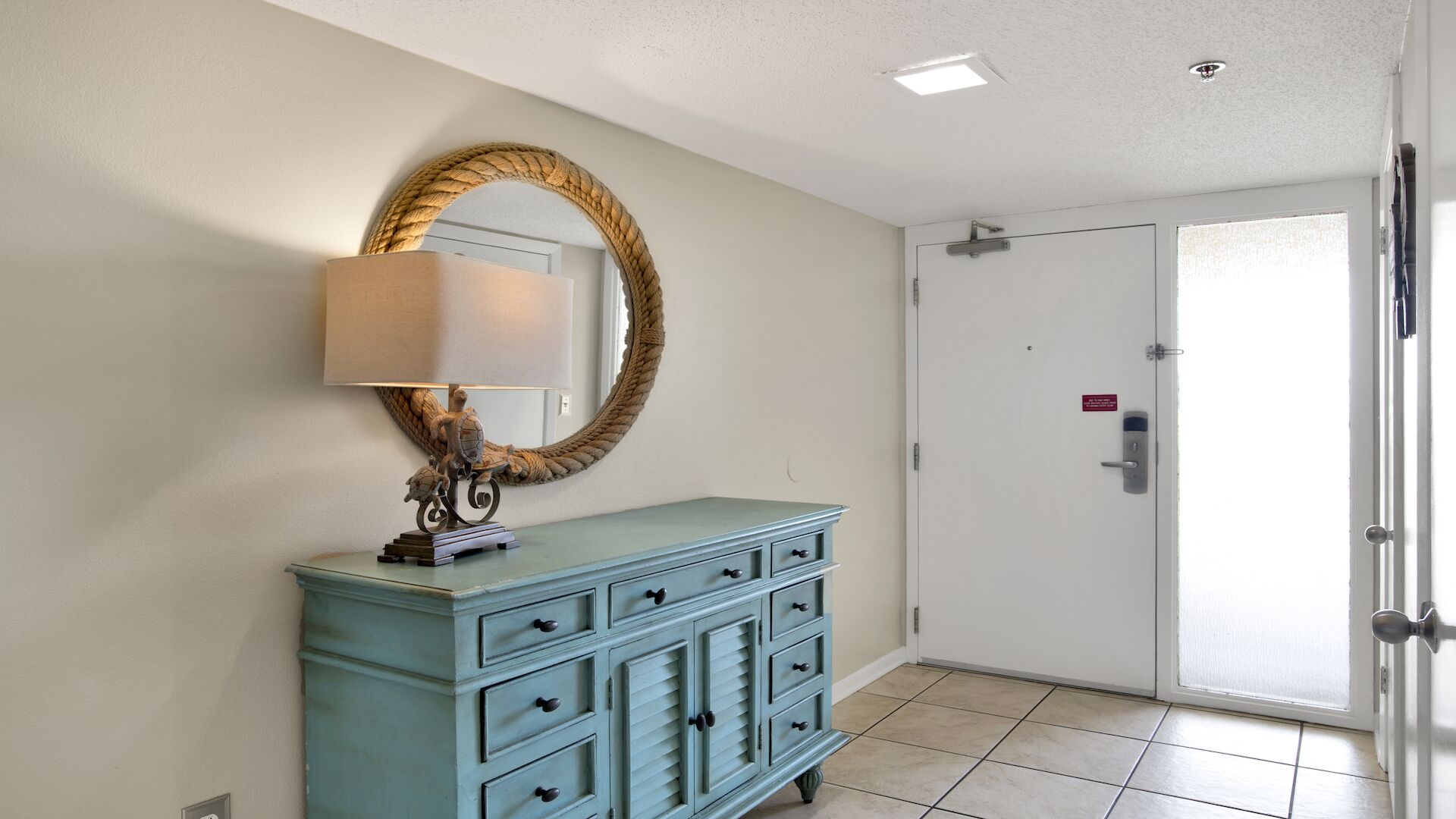 Un couloir avec une commode bleu clair, un miroir rond encadré d’une corde et une porte blanche dotée d’un panneau en verre dépoli.