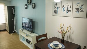 Een moderne woonkamer met een flatscreen televisie op een witte standaard, een eettafel met een bloevas en abstracte wanddecoratie.