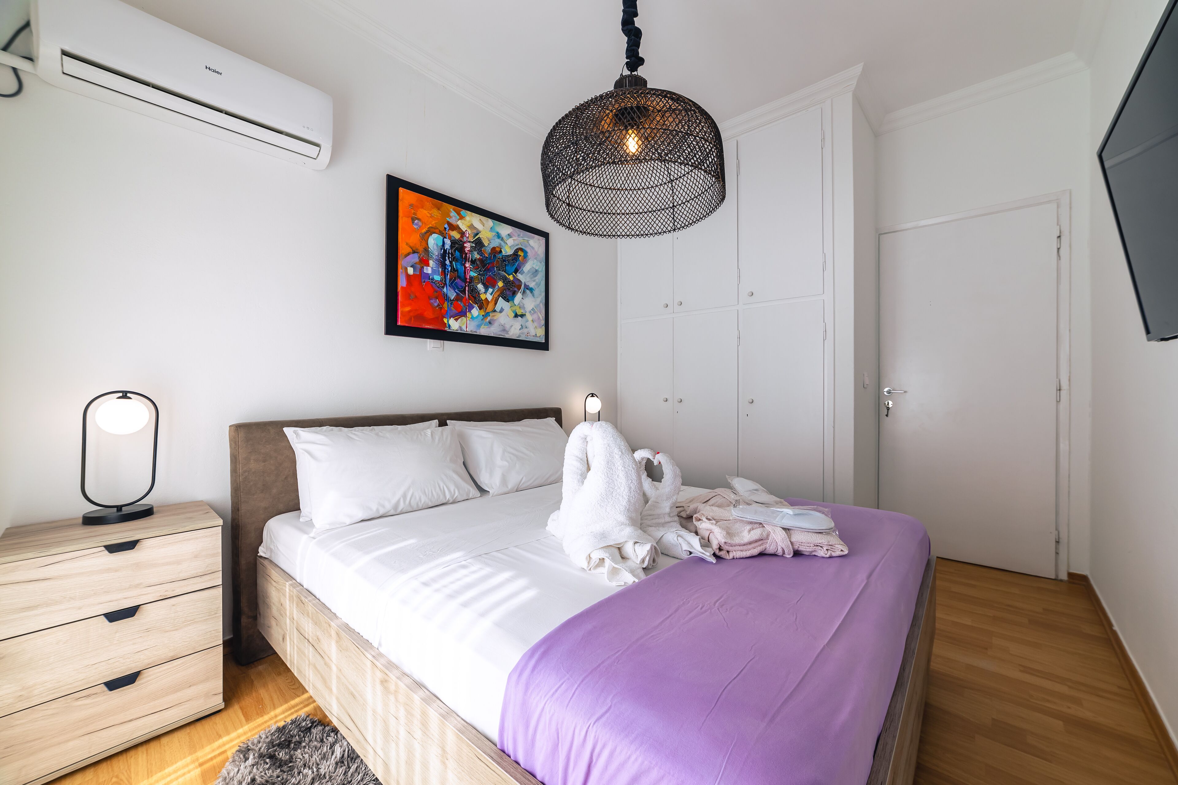 A neatly arranged bedroom with a bed, a bedside table, a lamp, an air conditioner, a colorful painting, and a hanging light fixture.