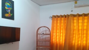 Una habitación con televisión montada en la pared, un estante de madera, una cortina naranja y una cama con edredón rojo y blanco.