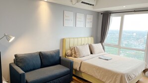 A modern bedroom with a bed, a sofa, a bedside table, and a window with a city view. - Breeze Apartments at Bintaro by OkeStay (South Tangerang)