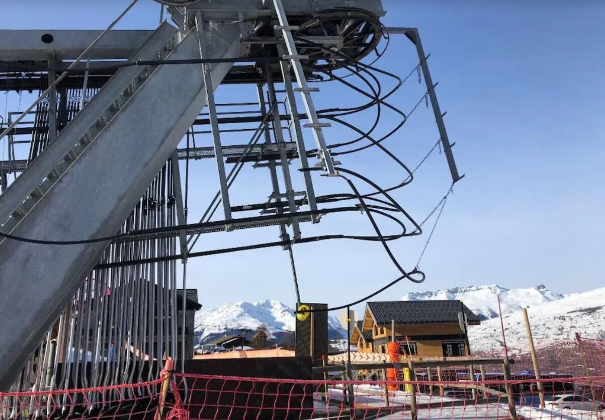 Een kabelbaanconstructie met kabels en een stoeltjeslift, met op de achtergrond besneeuwde bergen en een heldere lucht.