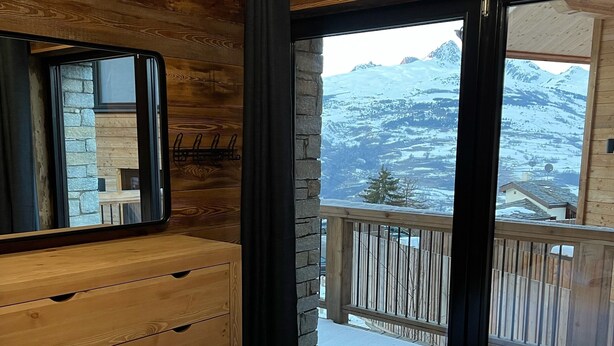 Un dormitorio con cómoda de madera, cama grande con ropa de cama blanca y vista a montañas nevadas desde un balcón.