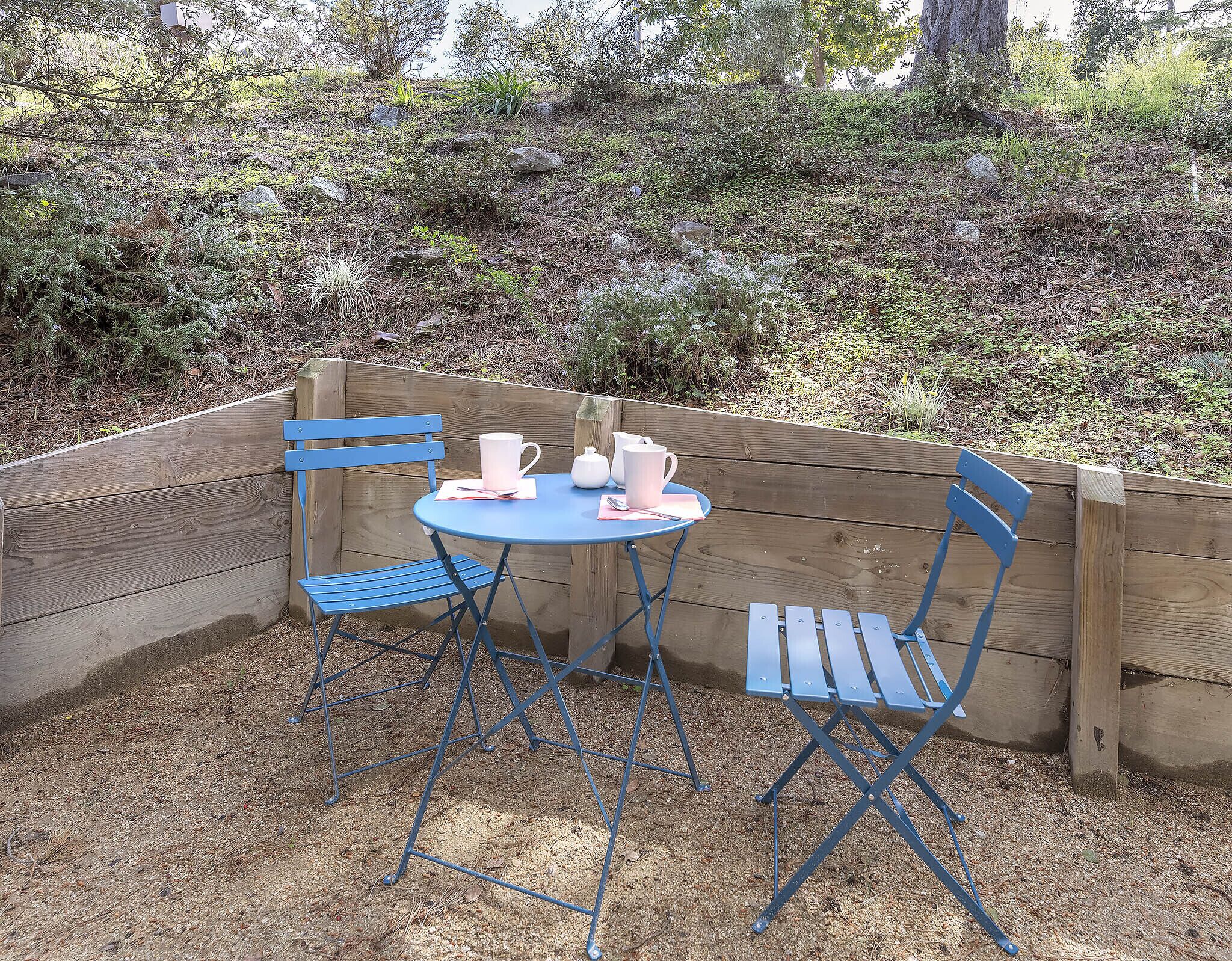 Une terrasse en bois avec une table bleue et des chaises assorties, un pichet blanc et une tasse posés sur la table.