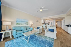 Un salon moderne avec un canapé bleu, des fauteuils blancs et une table basse. On peut voir un tableau au mur, un ventilateur de plafond et une vue sur la cuisine.