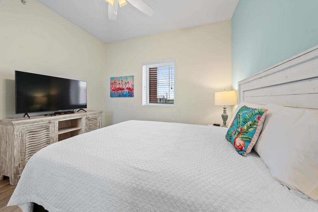 A bedroom with a large bed, a television on a wooden cabinet, a window with blinds, and a painting on the wall.
