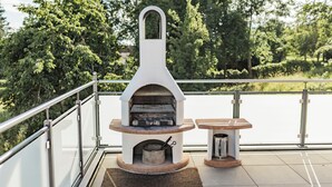 An outdoor grill with a stone base and a metal chimney, accompanied by a small table and a trash can, set against a backdrop of trees and a railing. - Ferienhof Hofer (Absberg)