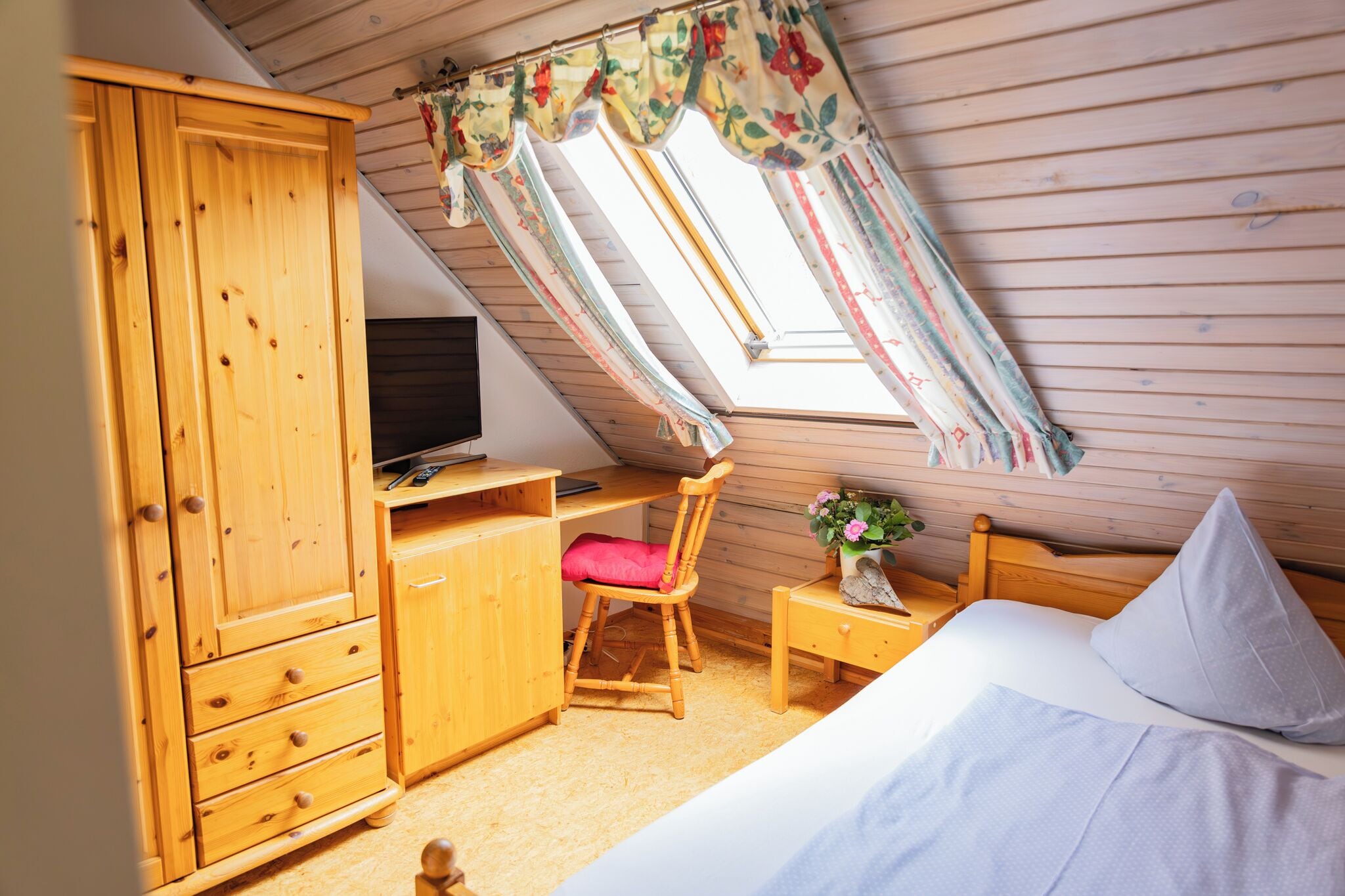 Un dormitorio con un armario de madera, un escritorio con silla, una cama con almohadas y un tragaluz con cortinas.