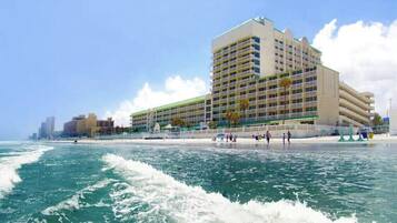 Un hôtel à plusieurs étages, doté de balcons, situé près d’une plage où l’on peut voir les vagues.