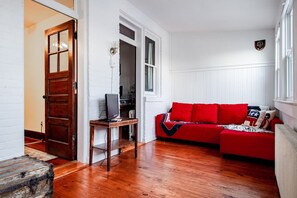 Ein Zimmer mit Holzboden, einem roten Sofa mit Kissen, einem Holztisch mit einem Computer und einer Tür zu einem anderen Raum.