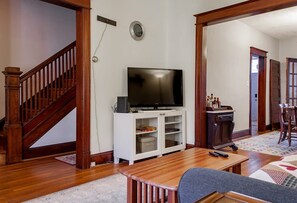 Un salon avec un escalier en bois, un téléviseur à écran plat posé sur un meuble blanc, un coin repas avec une table et des chaises, et un couloir menant à une autre pièce.