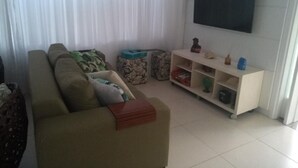 Un salon avec un canapé, un téléviseur fixé au mur, un meuble blanc et une fenêtre avec des rideaux.