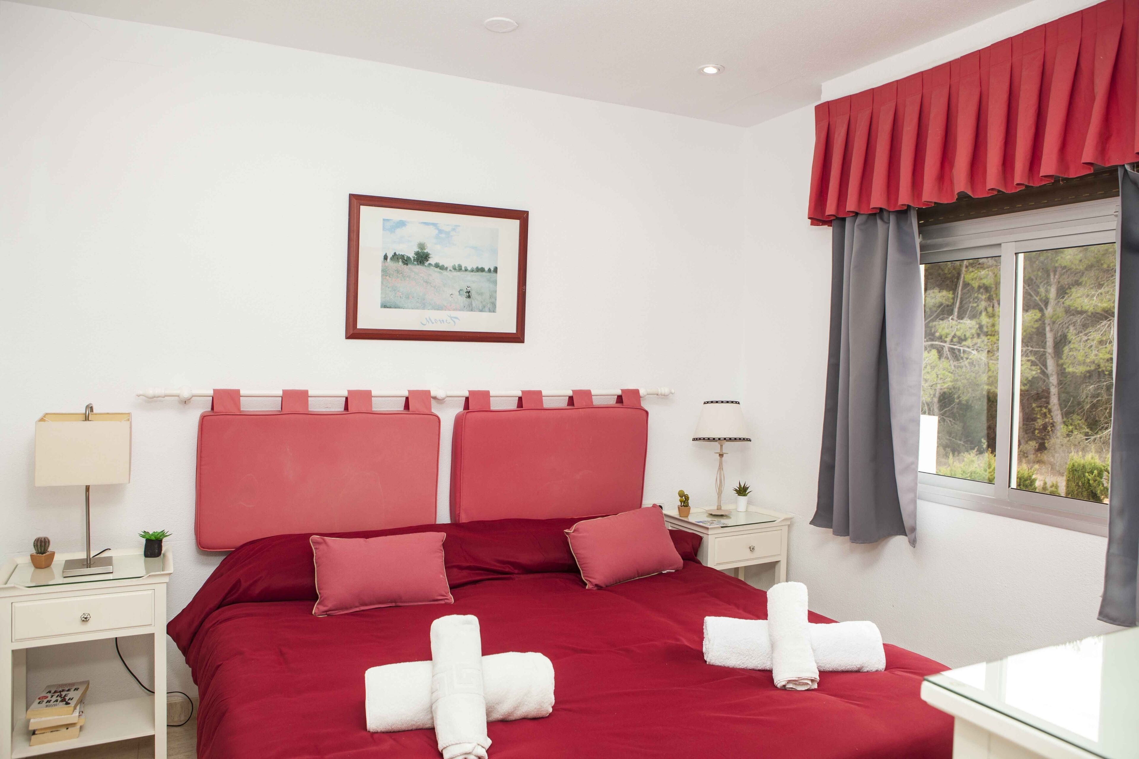 A neatly arranged bedroom with a red bedspread, a framed picture on the wall, and a window with curtains.