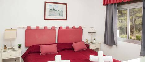 A neatly arranged bedroom with a red bedspread, a framed picture on the wall, and a window with curtains.