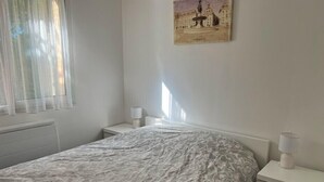 Un dormitori amb un llit, dues taules de nit, una finestra amb cortines translúcides i un quadre penjat a la pared que representa una font.