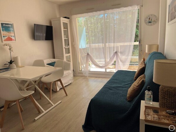 A room with a white table, chairs, a blue sofa, a TV on the wall, and a window with a view of greenery.