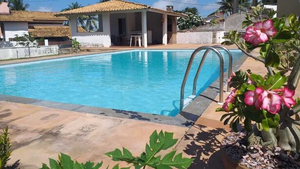 Una piscina con solárium empedrado, una zona de estar cubierta y una maceta con flores rosadas.