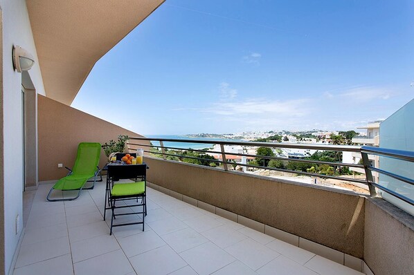 A balcony with a green chair, a table with a pitcher of orange juice, and a view of the cityscape.