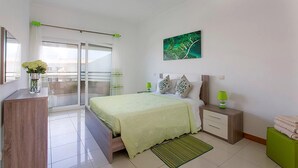 A modern bedroom with a bed, bedside tables, a green rug, a painting, and a balcony with a view. - Albufeira Beach Apartment with sea views of the Bay all the way to the Old Town (Albufeira)