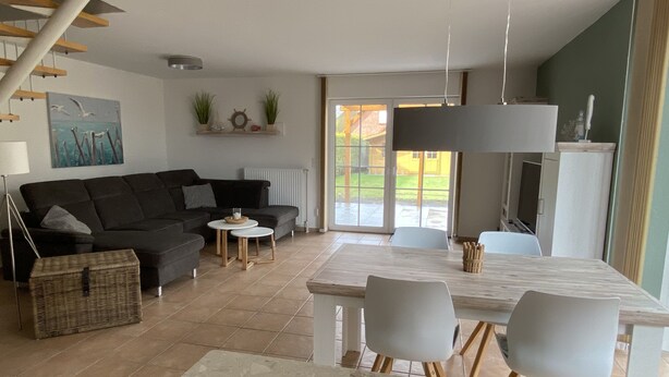 Een moderne woonkamer met een donkerbank, een klein rond tafeltje, en een eethoek met een witte tafel en stoelen.