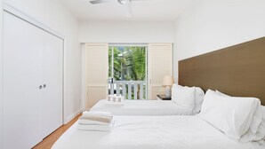 A bedroom with two beds, a ceiling fan, a nightstand with a lamp, and a view of greenery through a door.