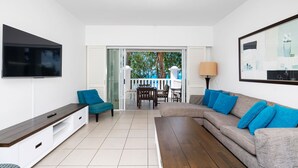 A living room with a TV, sofa, and coffee table. There is a balcony with a view of trees and a white fence.