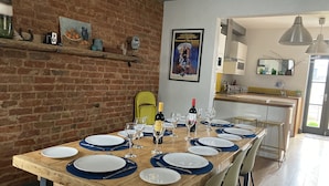 Uma sala de jantar com uma longa mesa de madeira posta para seis, uma parede de tijolos e uma cozinha ao fundo.
