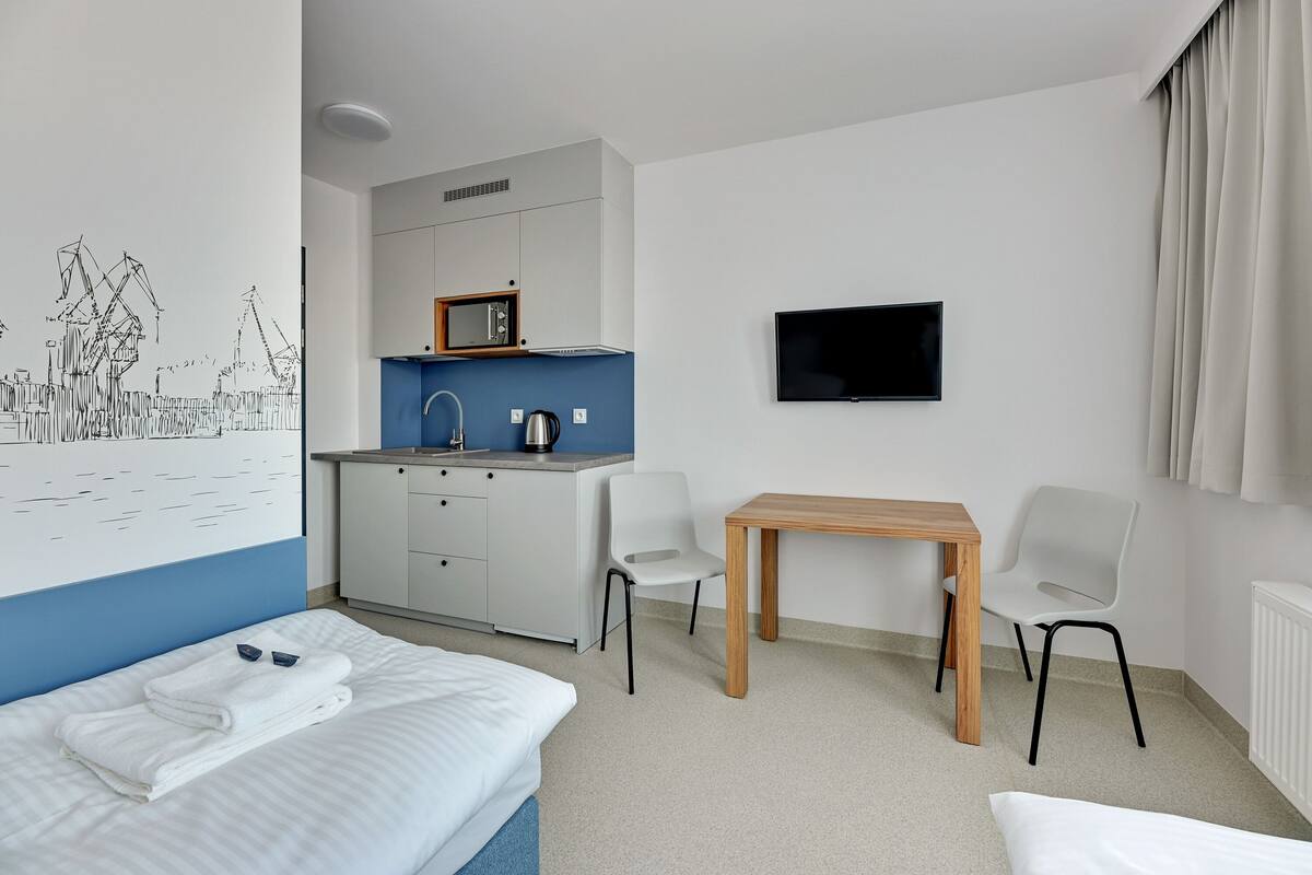 Ein modernes Hotelzimmer mit Bett, Schreibtisch, Stuhl, Fernseher und einer Küchenzeile.