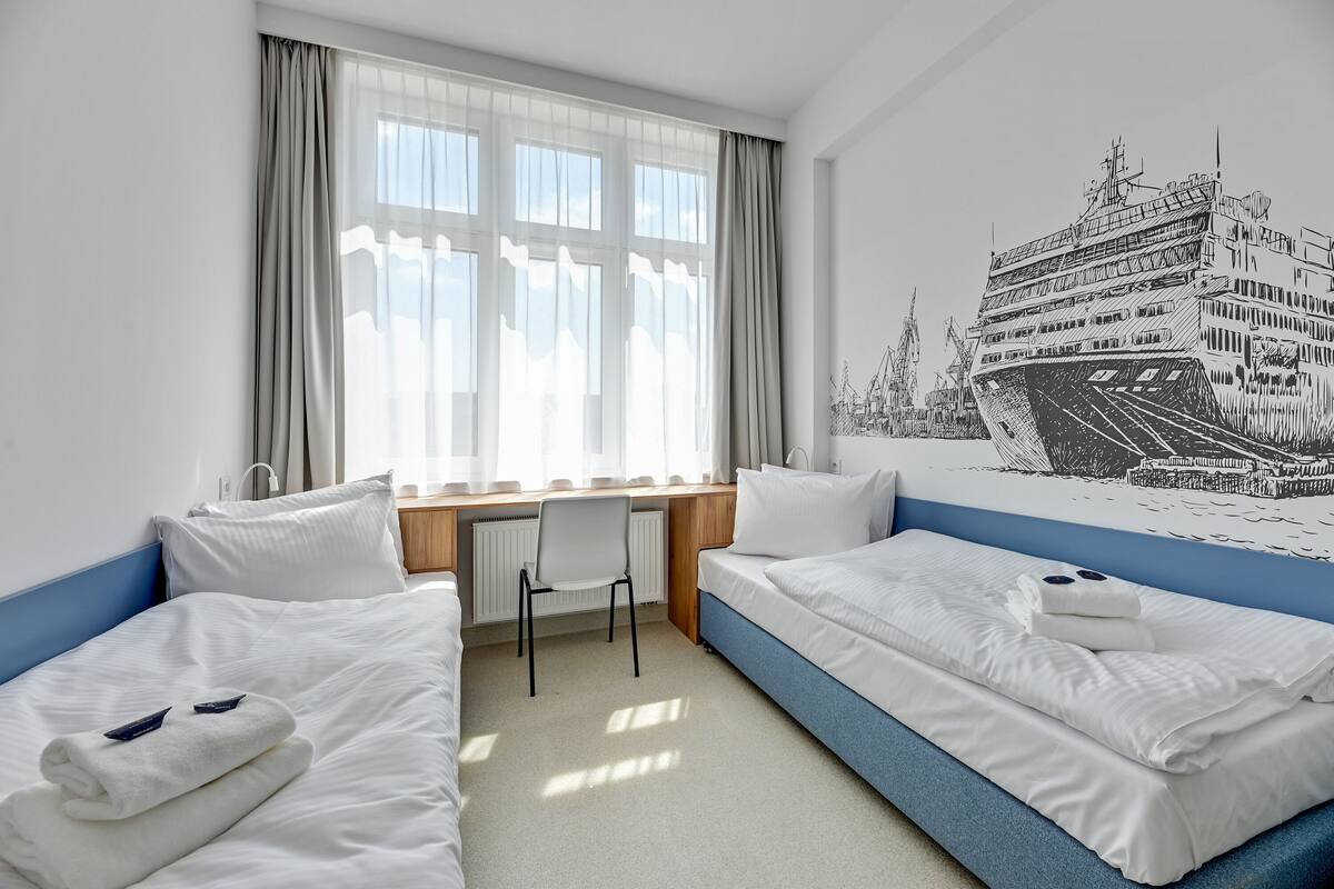Ein Hotelzimmer mit zwei Betten, einem Schreibtisch und einem großen Fenster mit Vorhängen.