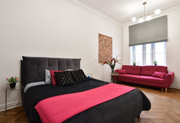 A bedroom with a large bed, a red sofa, a brick accent wall, and a window with blinds.
