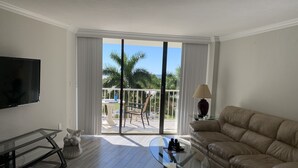 A living room with a sofa, a glass coffee table, a TV, and a balcony with a view of palm trees. - Updated unit with sunset & beach view (Marco Island)