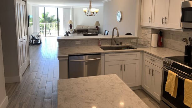 A modern kitchen with white cabinets, a stainless steel dishwasher, and an oven. There is a large window providing natural light and a view of the outdoors. - Updated unit with sunset & beach view (Marco Island)