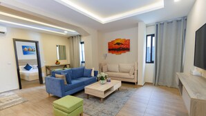 Een moderne woonkamer met een blauwe bank, een beige bank, een houten salontafel en een groene poef. Er hangt een schilderij aan de muur en bevindt zich een televisie aan de rechterkant.