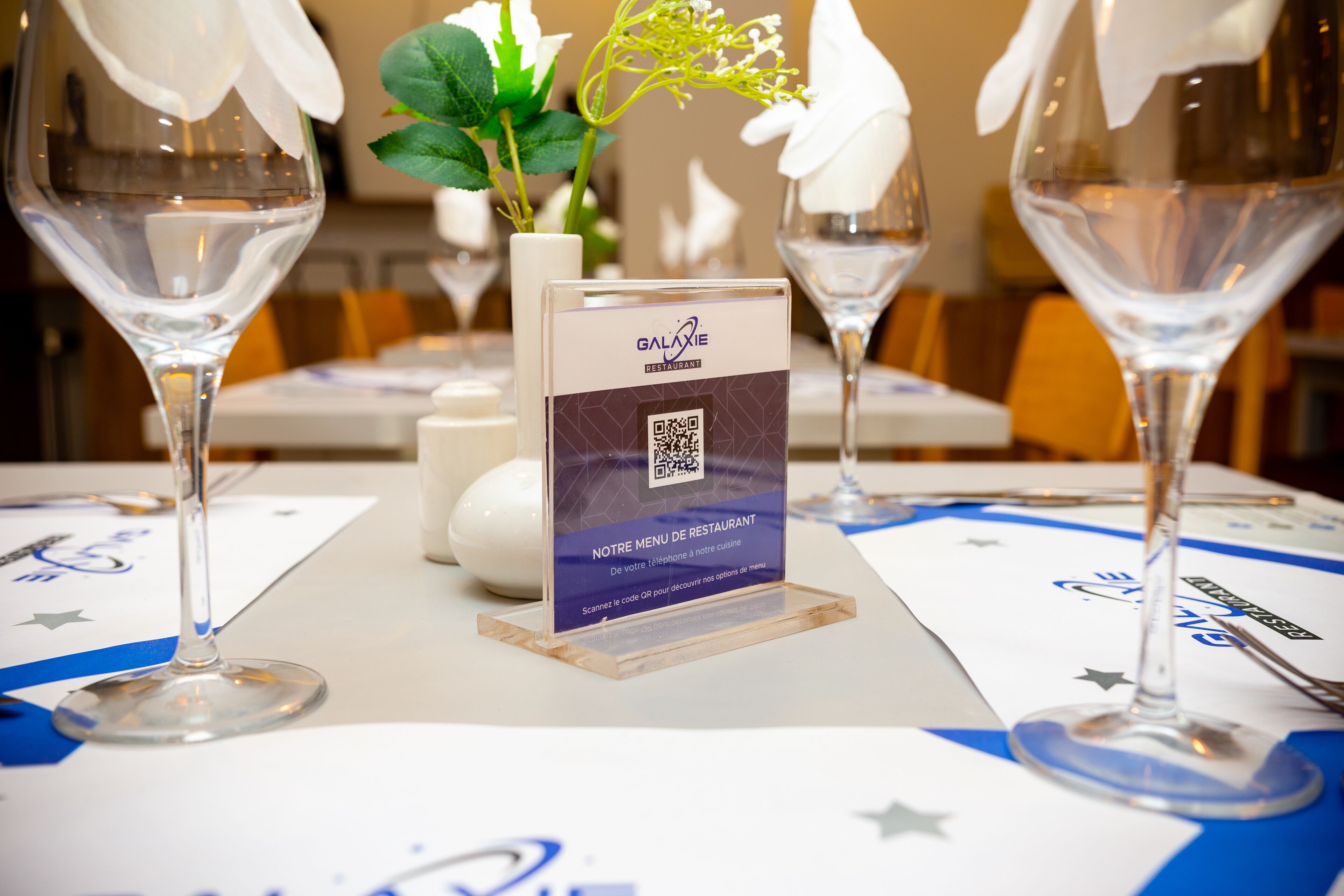Une table de restaurant avec un présentoir à code QR pour afficher le menu, des verres à vin et un vase rempli de fleurs.