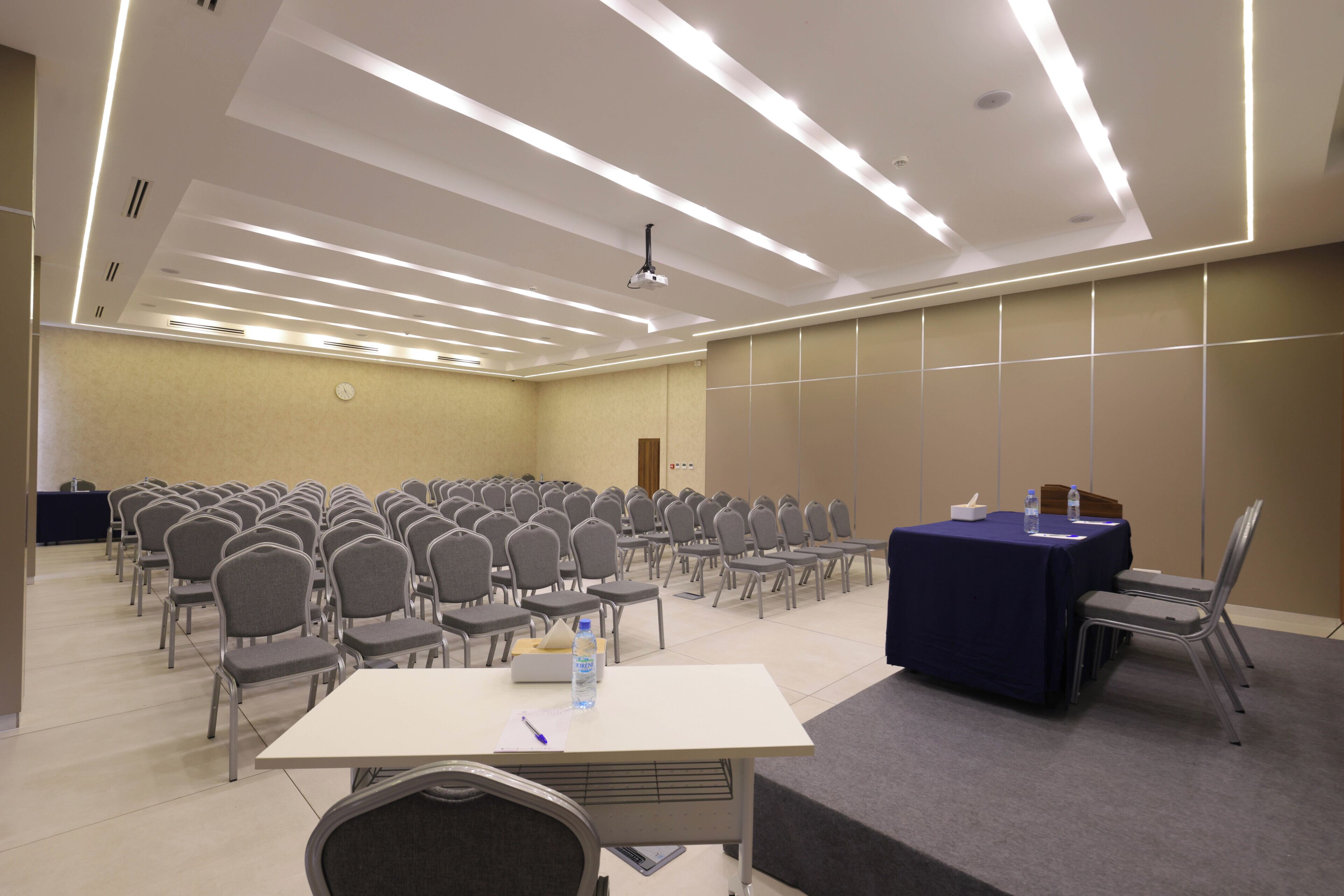 Un salón de conferencias con hileras de sillas, una mesa con mantel azul y un proyector en el techo.