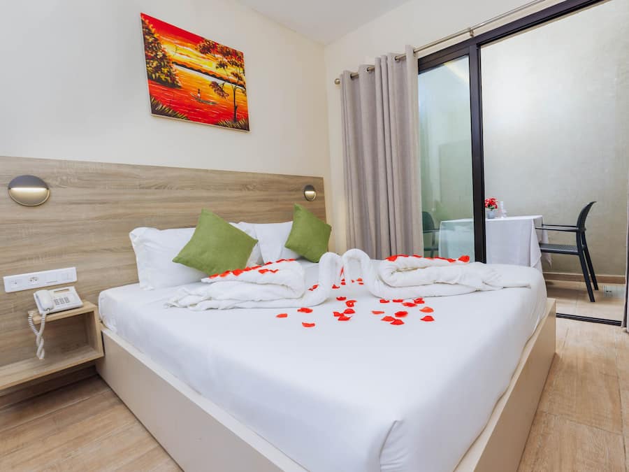 Una cama bien hecha con sábanas blancas, almohadas verdes y pétalos de rosa roja. Hay un cuadro en la pared, un teléfono en la mesita de noche y una silla junto a la ventana.