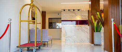 A hotel reception area with a marble reception desk, a gold reception stand, a red carpet, and a seating area with a grey sofa.