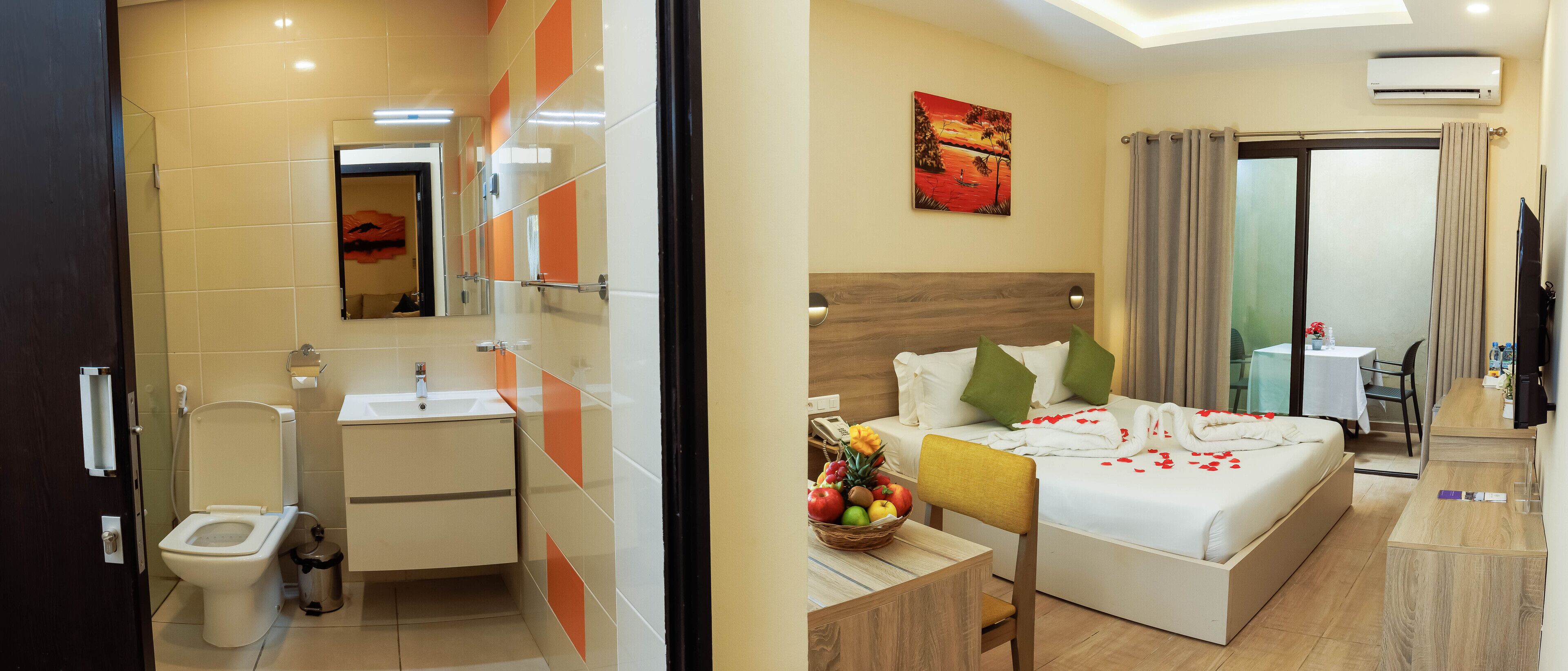A hotel room with a bed, a bedside table with a fruit basket, a bathroom with a toilet and sink, and a TV.